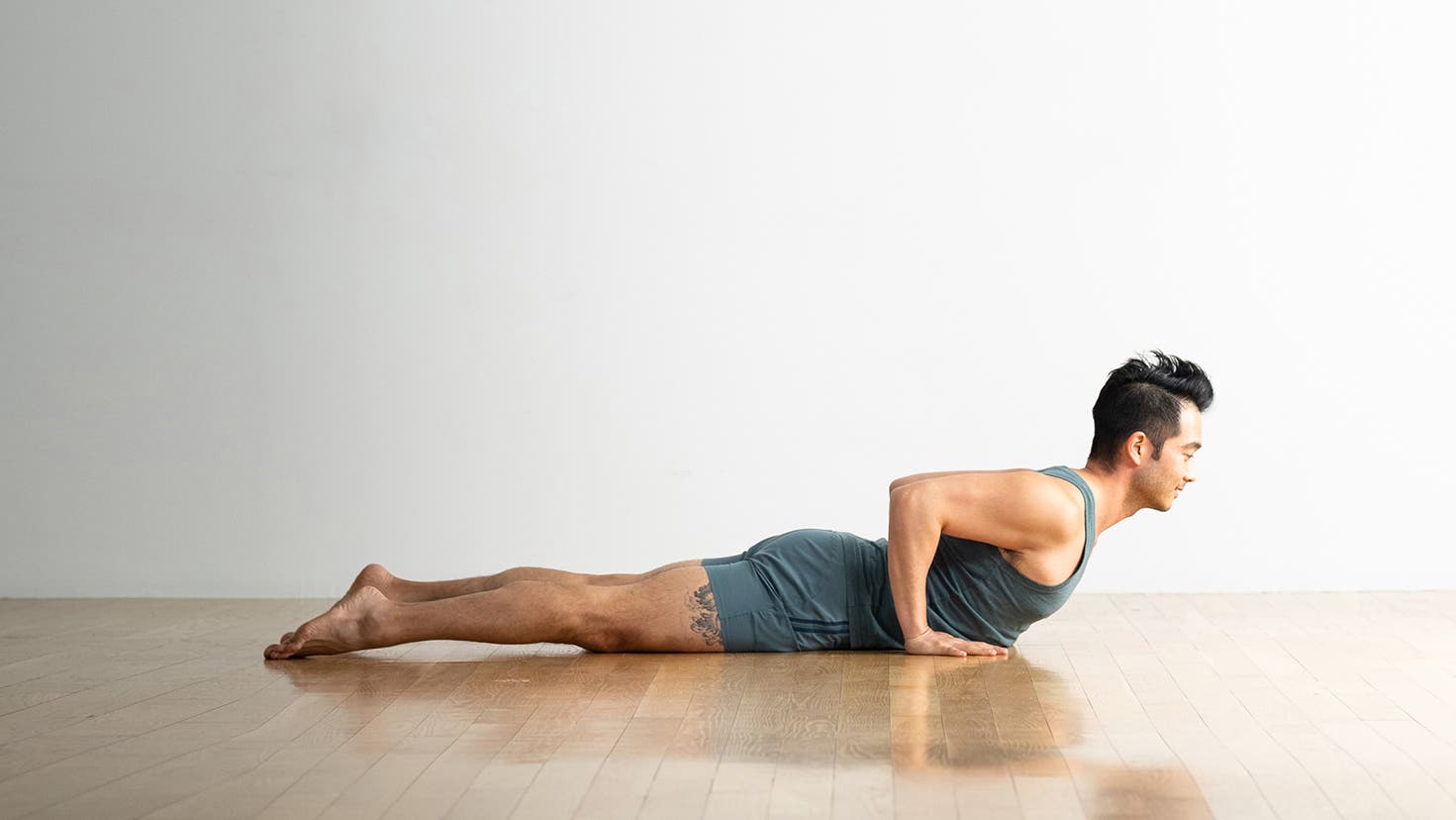 Hiro Landazuri in blue-gray shorts and top is lying on a wood floor, practicing Cobra Pose