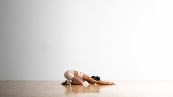 A woman practices Extended Child's Pose (Balasana) on a wood floor against a white background