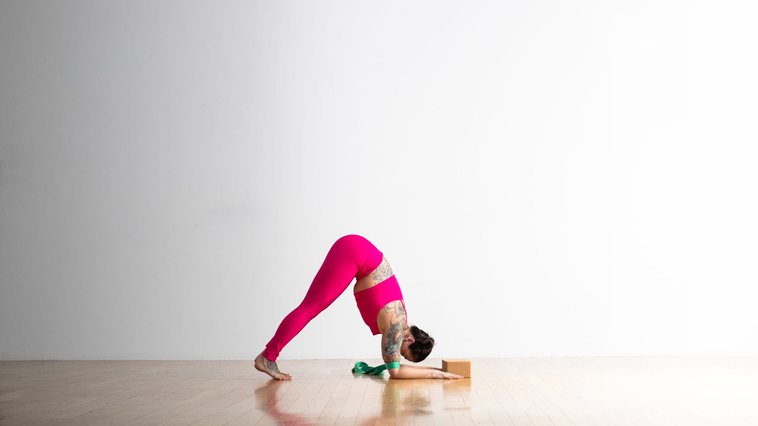 A woman practices Dolphin Pose with a green strap around her upper arms. A cork block is placed between her hands. She is wearing bright magenta yoga tights and a matching crop top.