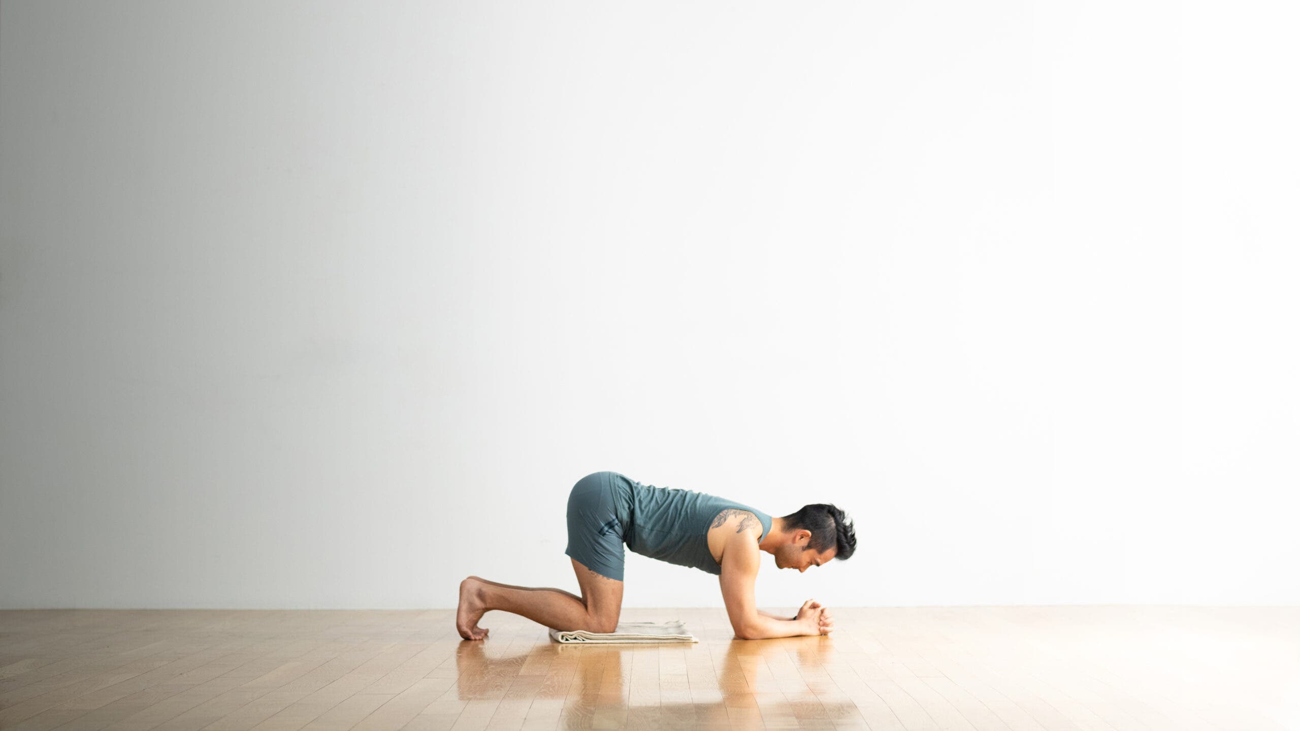 A man kneels in a variation of Tabletop Pose in preparation for Dolphin Pose. He is kneeling on a blanket and has his forearms on the floor. He's wearing blue shorts and a top.