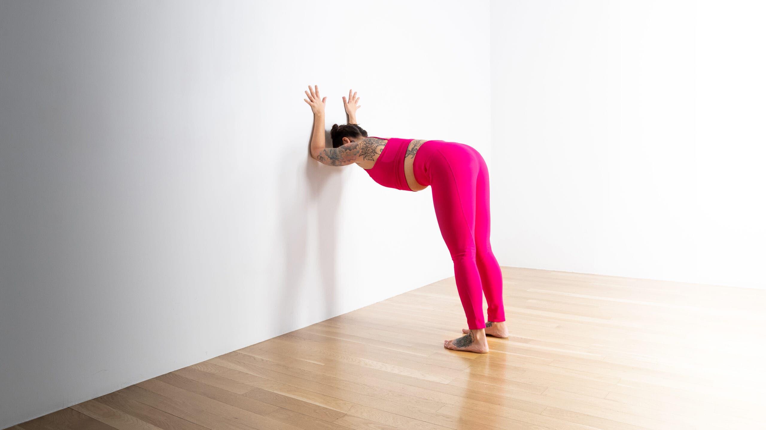 A woman practices a variation of Dolphin Pose at the wall. She is standing a few feet from a white wall, bends at the hips and brings her forearms to press against the wall. She is wearing bright magenta yoga tights and a matching crop top. She has dark hair in two buns and has tattoos on her feet back and arms.
