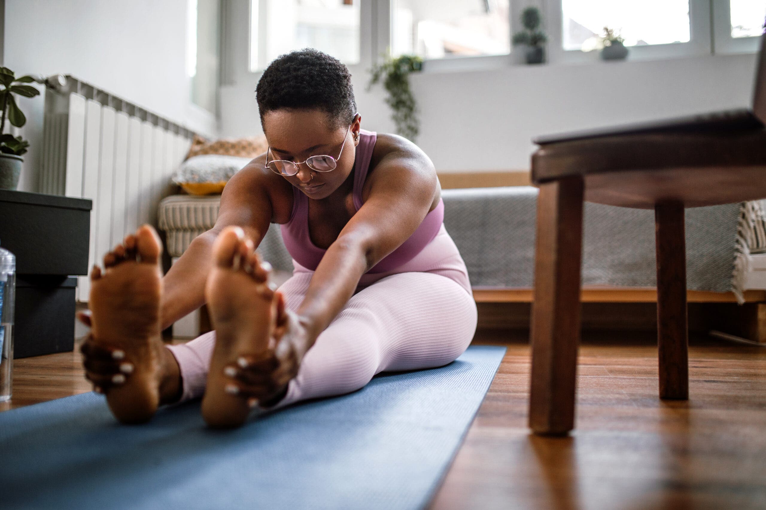 A brown-skinned woman with short cropped hair reaches forward to grasp her feet in Paschimottanasana (Seated Forward Bend). She is wearing a dark mauve top and pink yoga tights. The floor is dark wood and her yoga mat is blue. The walls are white and sunlight shines through uncovered windows. There is a sofa or bed behind her and a wooden chair or bench is to her left.
