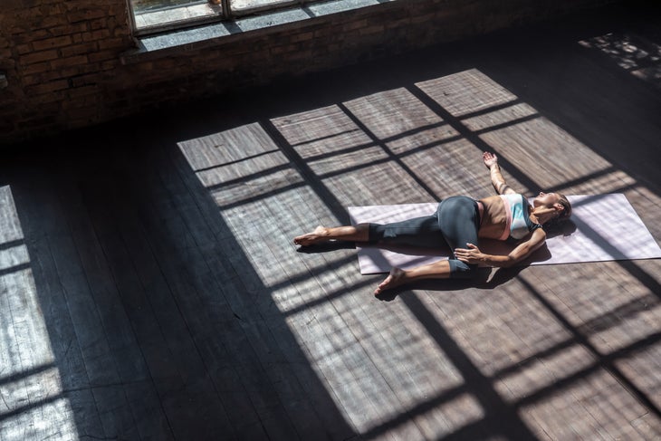 Woman doing a reclining twist on a gray floor. Shadows cast by the windows darken the room.