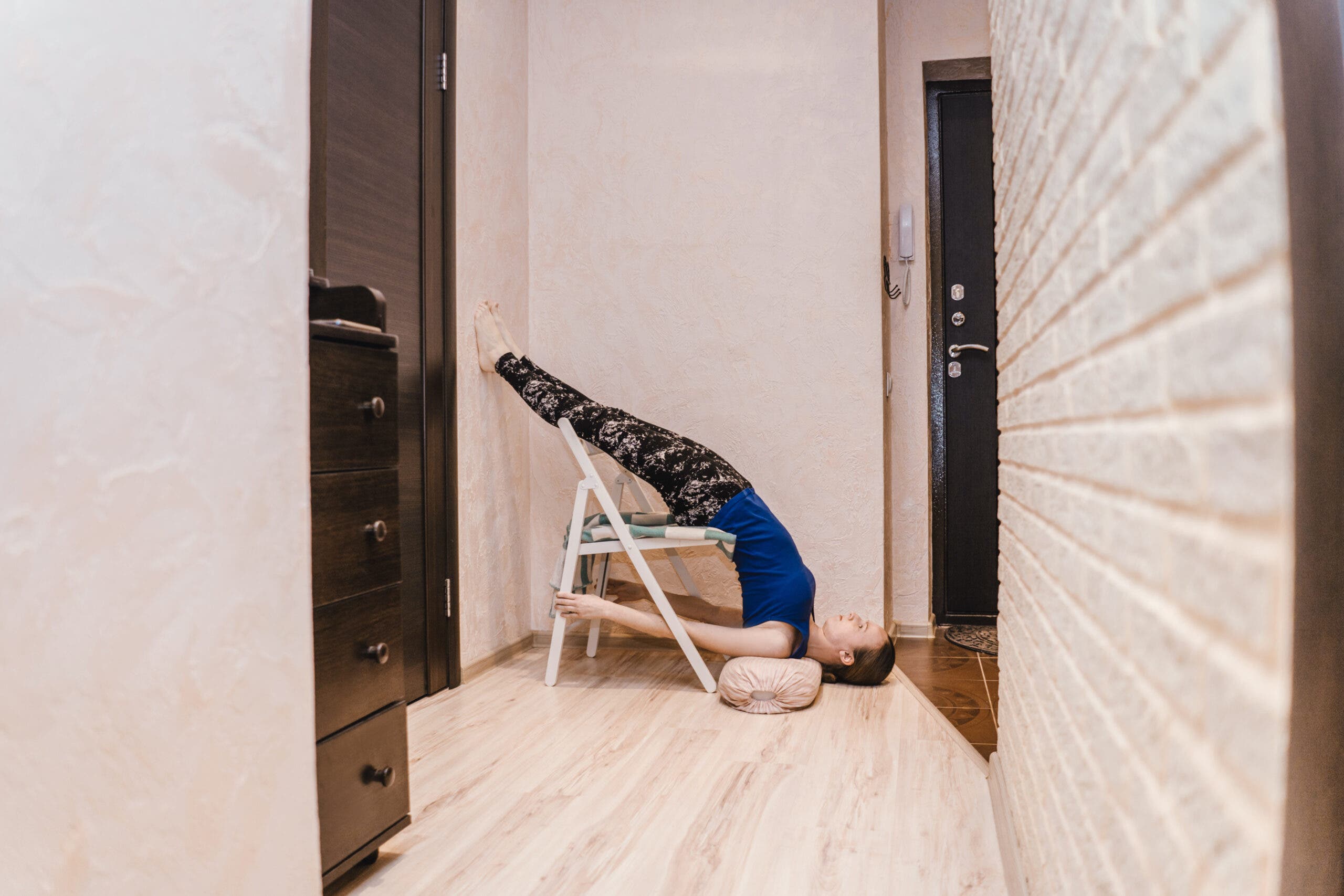 Woman in blue shirt and patterned yoga pants, practices a shoulder stand using a chair as a prop. She is in a narrow hallway with white brick walls and black doors. 
