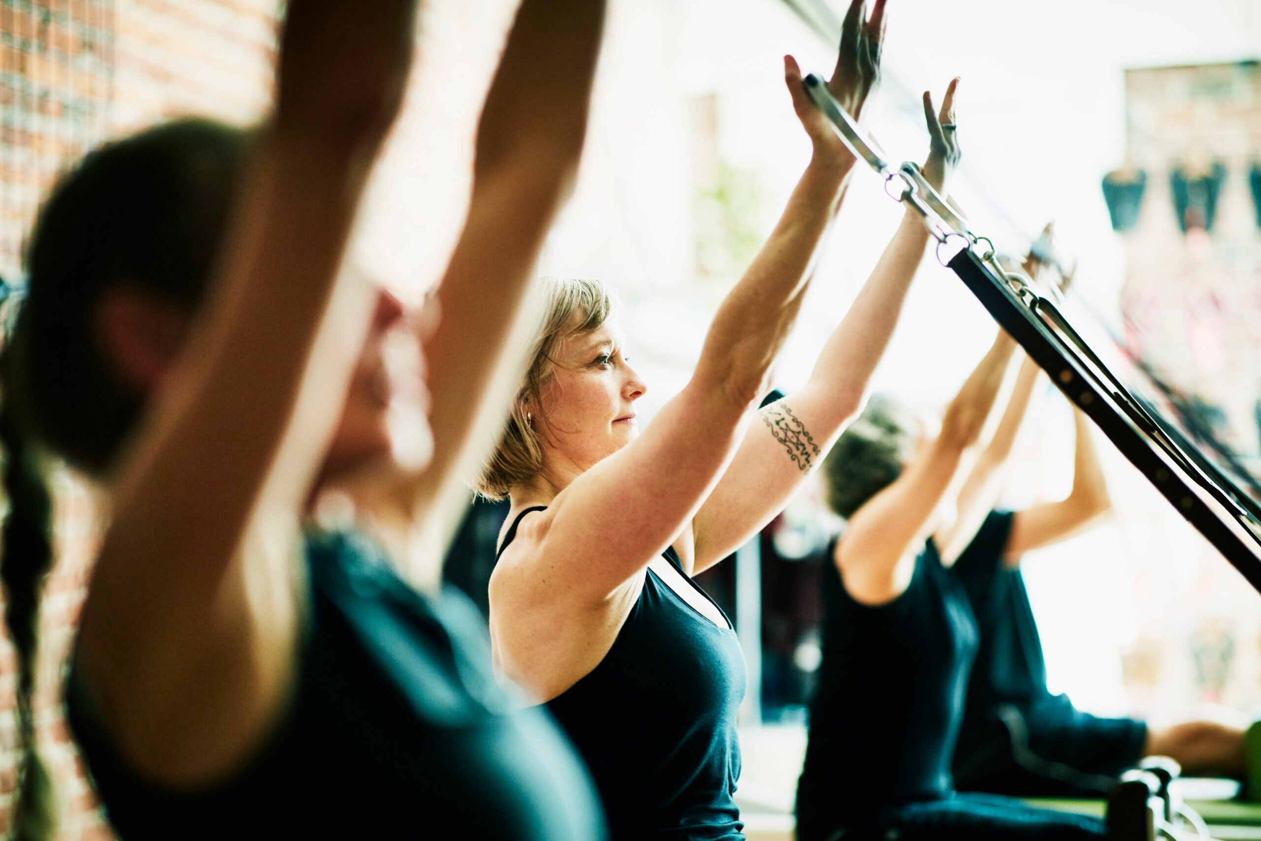 Women using reformer arm straps during pilates class in fitness studio.