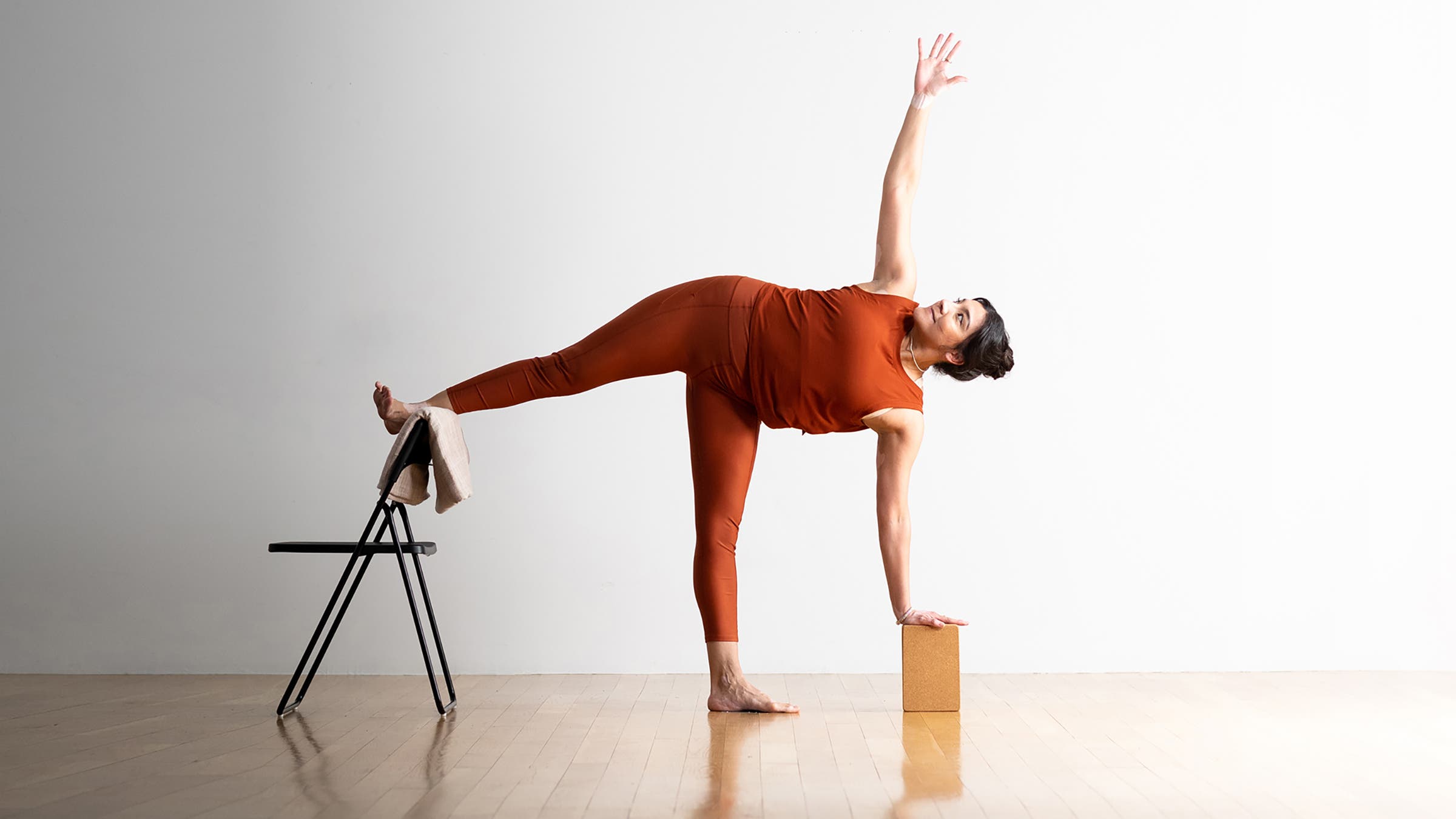 A person demonstrates a variation of Half Moon Pose in yoga, with one hand on a block and another on a chair