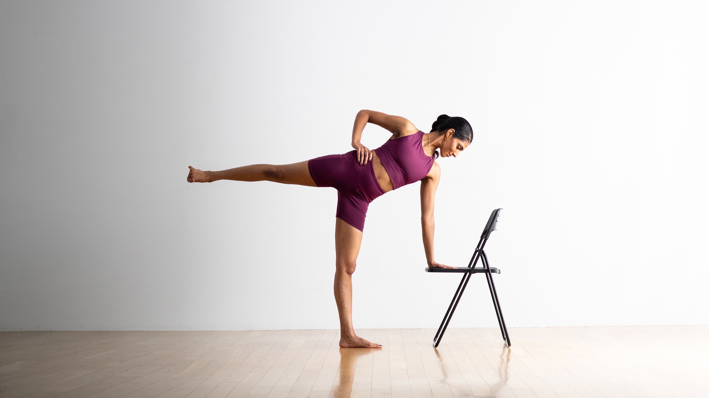 A South Asian woman wearing burgundy shorts and tank practices Half Moon using a folding chair as a support