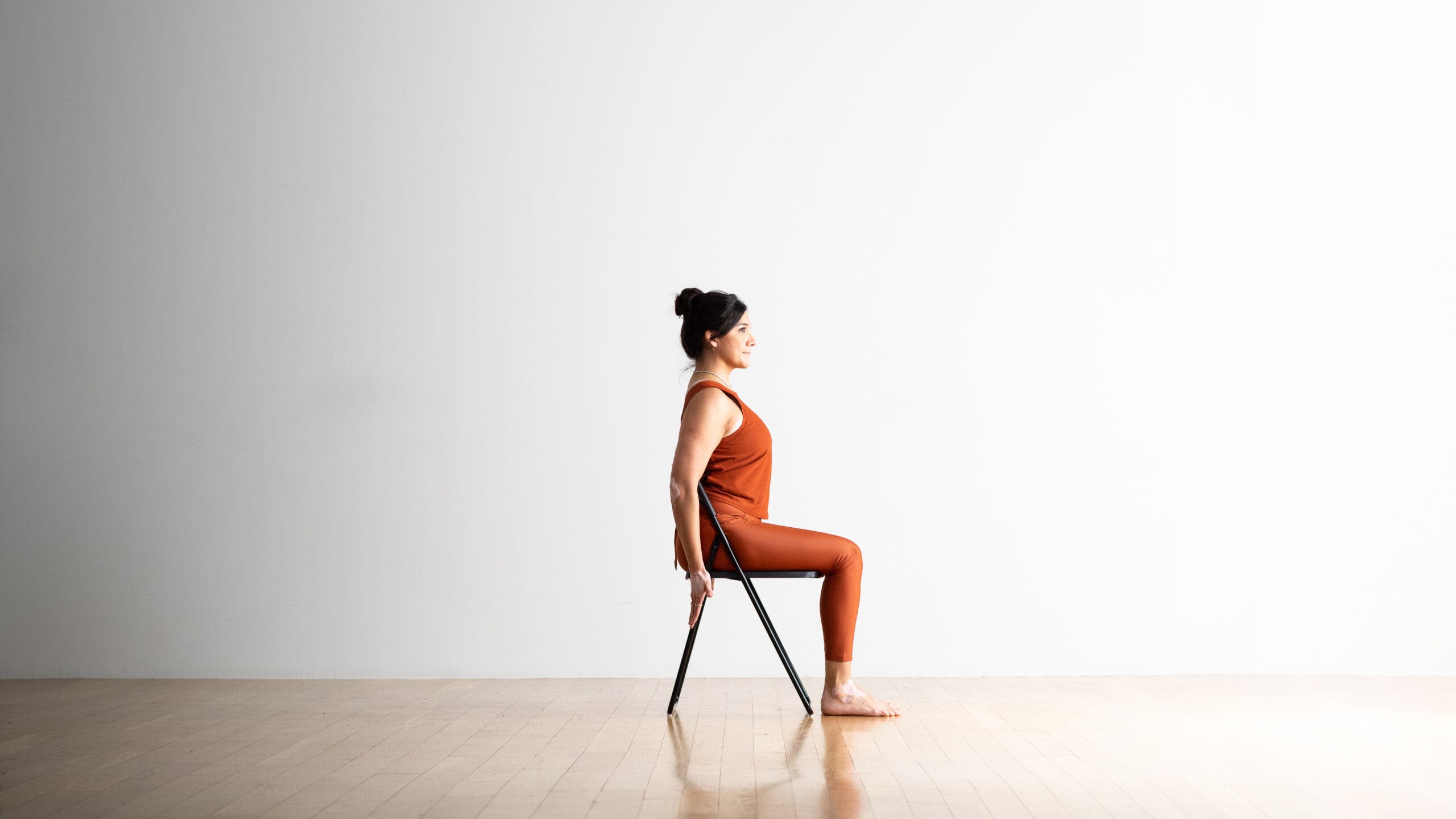 A woman with dark hair sits in a chair with her feet on the floor in a variation on Hero Pose. Her feet are on the floor and her hands are back along the back legs of the chair. She is wearing copper-colored yoga tights and a matching top. The floor is wood and there is a white wall in the background.