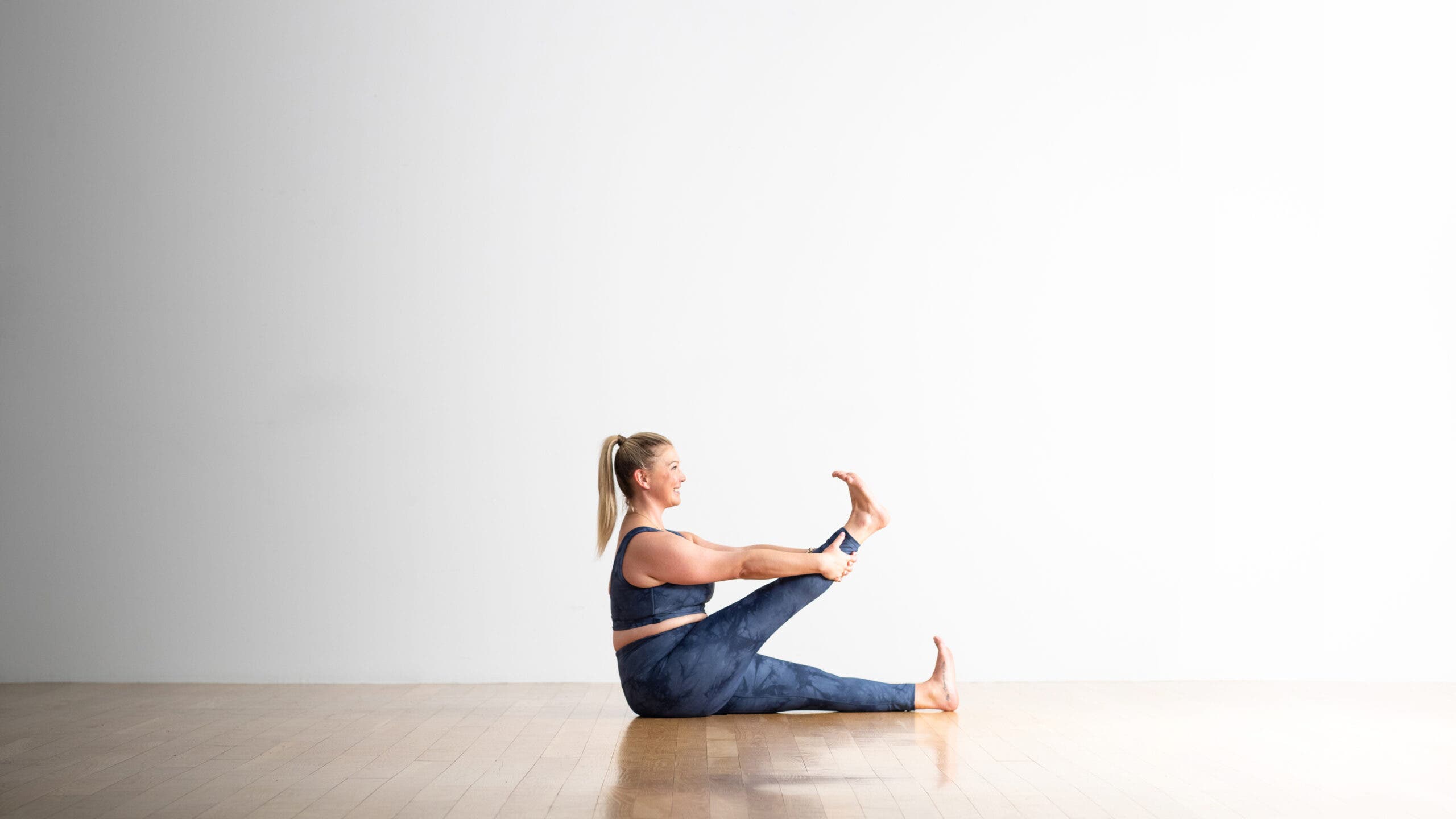 Sarra Raney practices Kraunchasana. She is sitting in Dandasana and lifts her right leg upholding it at the calf with both hands