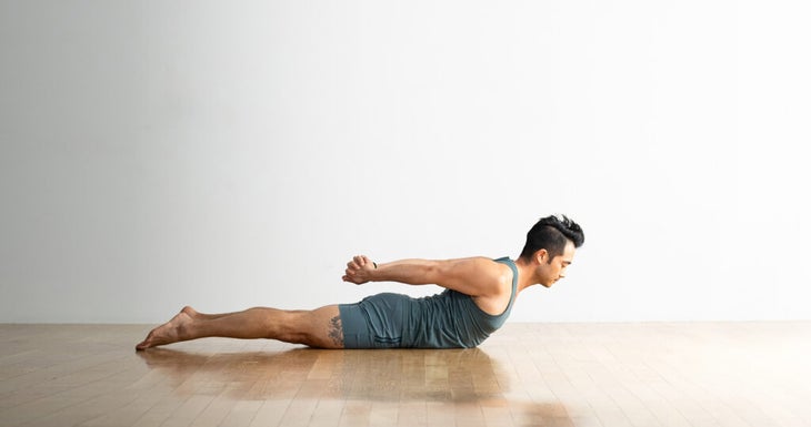 Hiro Landazuri übt die Half-Locust-Pose mit den Beinen auf dem Boden, den Schultern nach oben und den Händen hinter dem Rücken verschränkt