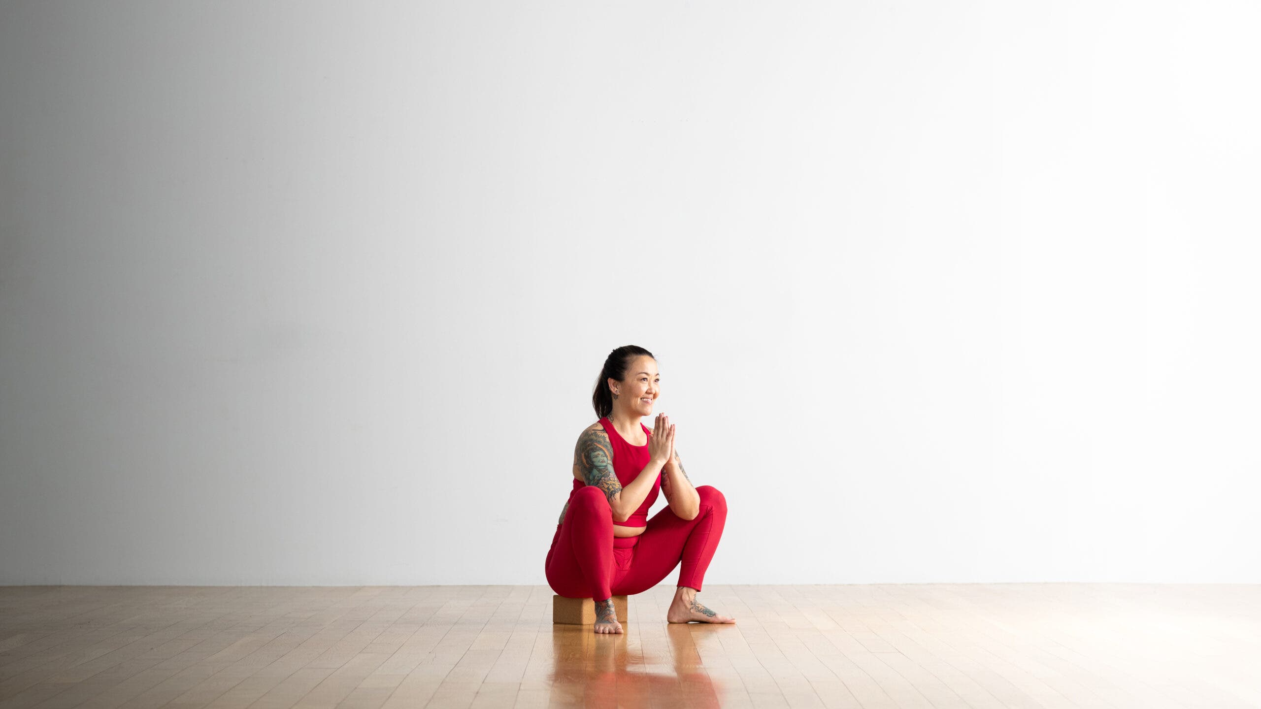 A woman practices Garland Pose with a block under her glutes. She had tatoos on her arm and foot. She is wearing bright pink yoga tights and a crop top. The room is white with a wood floor.