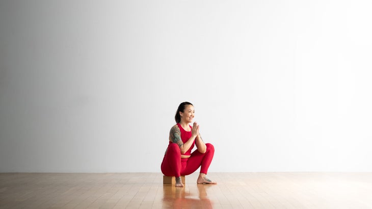 A woman practices Garland Pose with a block under her glutes. She had tatoos on her arm and foot. She is wearing bright pink yoga tights and a crop top. The room is white with a wood floor.