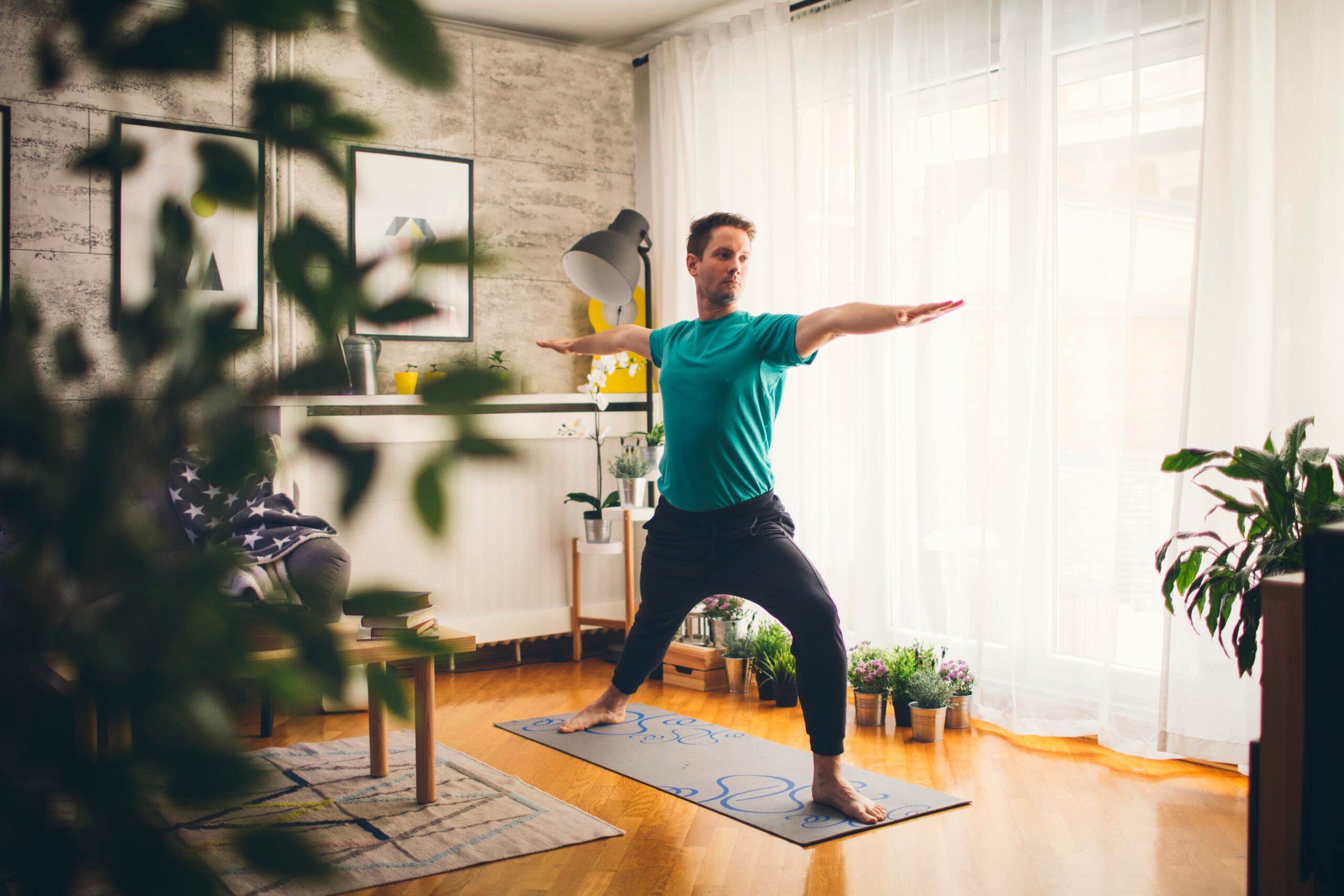 A man wearing a green shirt and black pants practices Warrior 2. His mat is gray with blue swirls and it set on a wood floor. At his back is a wall of windows covered by sheer white curtains. Small pots of plants are set on the floor and here are orchids on a plant stand. To his right is a wall that looks like large marble tiles with two abstract pieces of art. A white shelf under the paintings holds a yellow clock. There's a plant in the foreground at the left of the photo that partially obscures a chair with a star patterned blanket, a coffee table with books and an oatmeal colored woven rug.