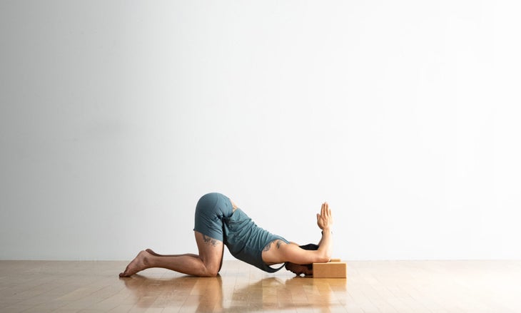 A man with dark hair practices Extended Puppy Pose with his elbows propped on cork blocks and hands in prayer position. He wears a blue-gray shorts and a top in the same color.