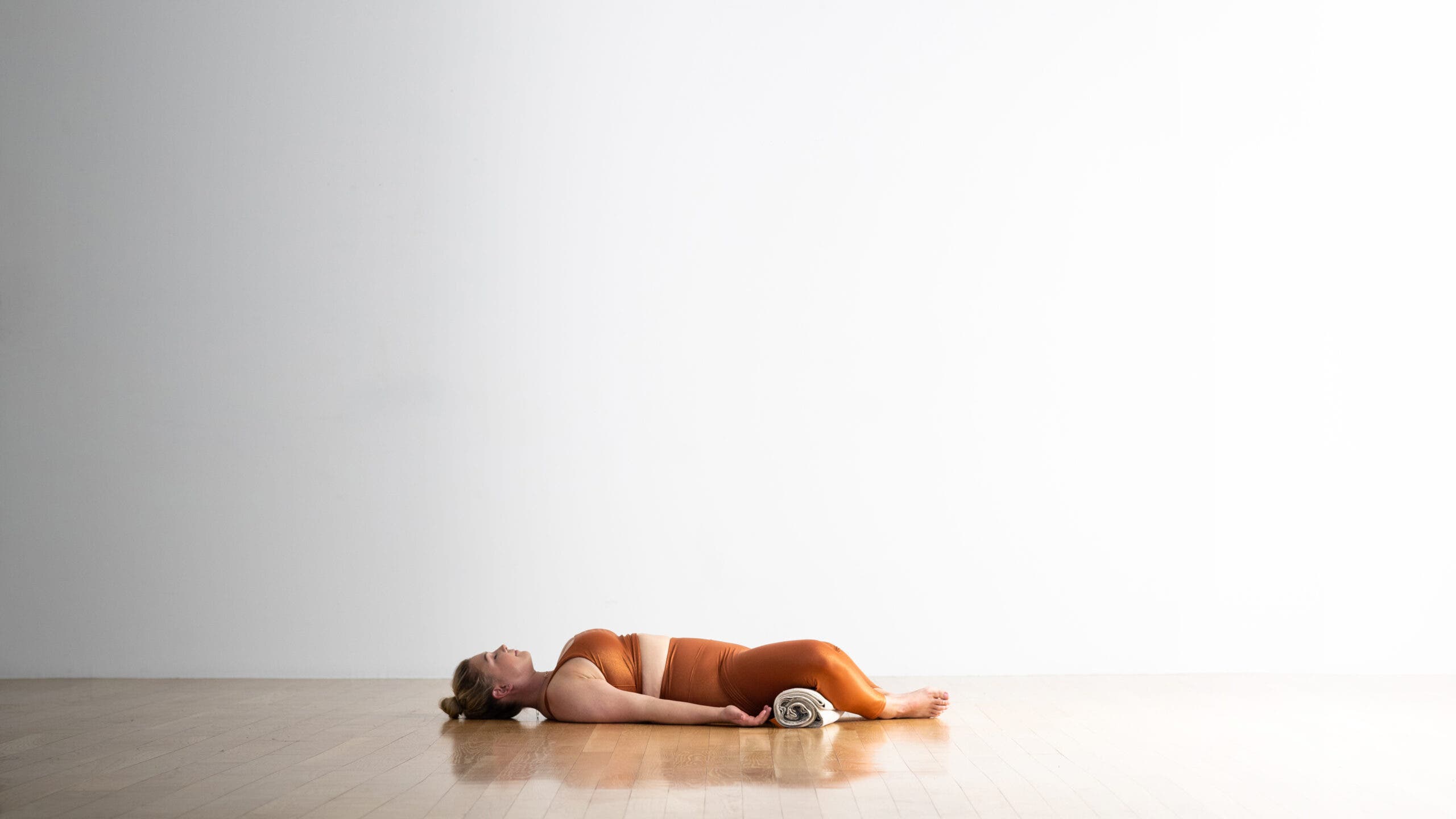 A woman practices Reclining Bound Angle Pose with a blanket roll under her knees. She is wearing shiny, golden yoga tights and a matching cropped top. Wood floor and white wall