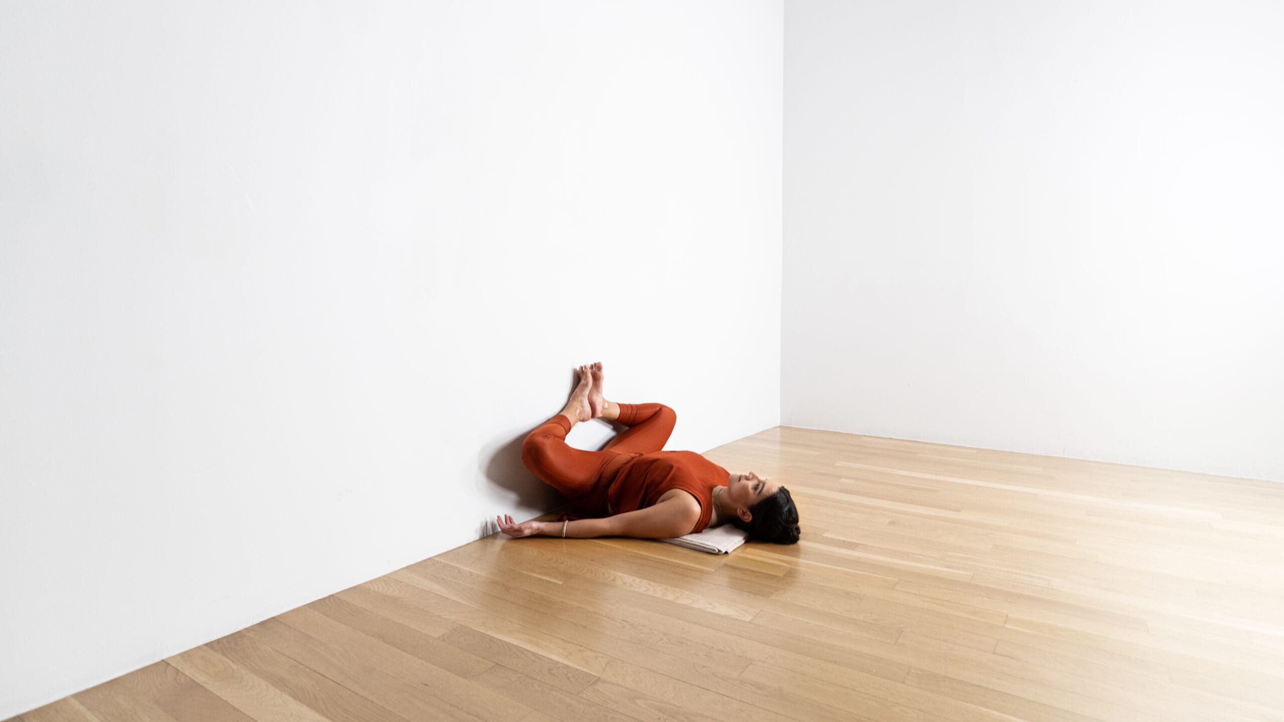 A woman practices Reclined Bound Angle pose against the wall. She is lying on a wood floor. Her legs against the wall, with her knees opened laterally.