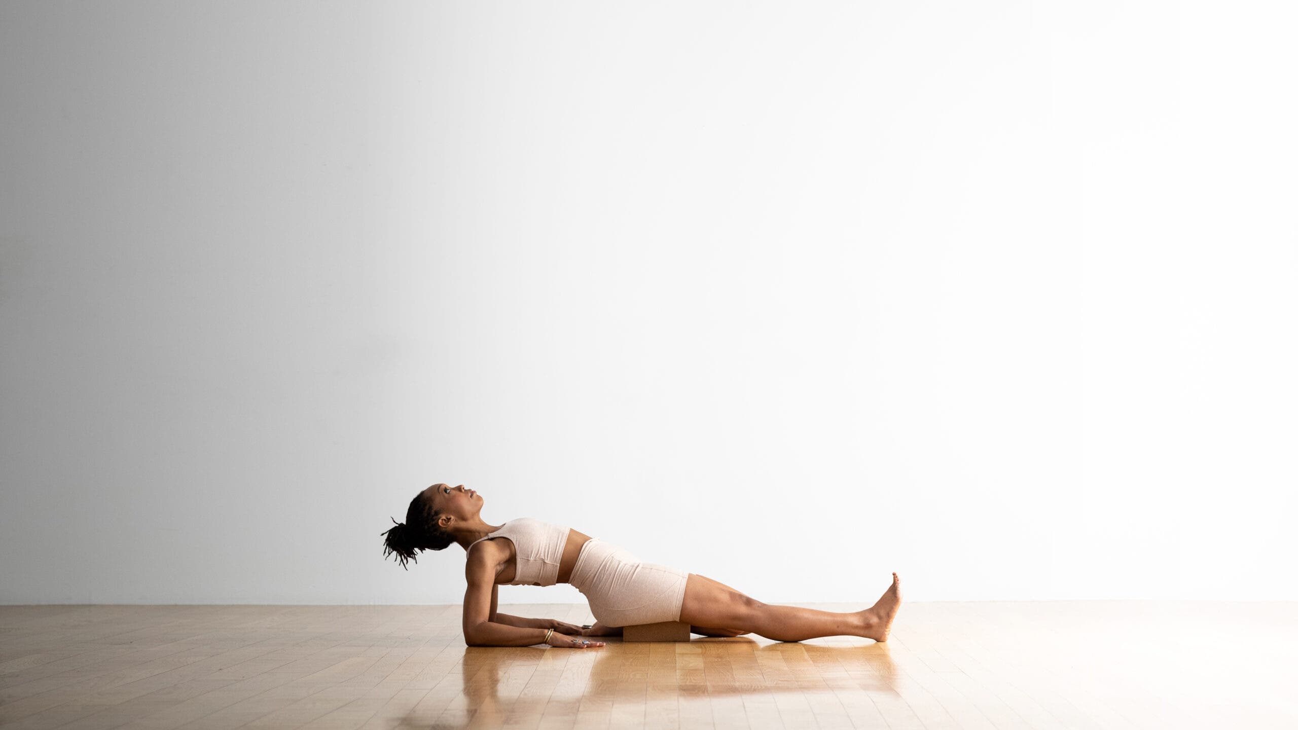 Woman practices Supta Ardha Virasana (Half Reclining Hero Pose. She draw just her right leg back into Virasana, and the other leg is extended. She leans back and props herself up on her elbows and forearms. She is a Black woman with a ponytail, wearing off whit yoga shorts and a matching top. The floor is wood and the wall behind her is white.