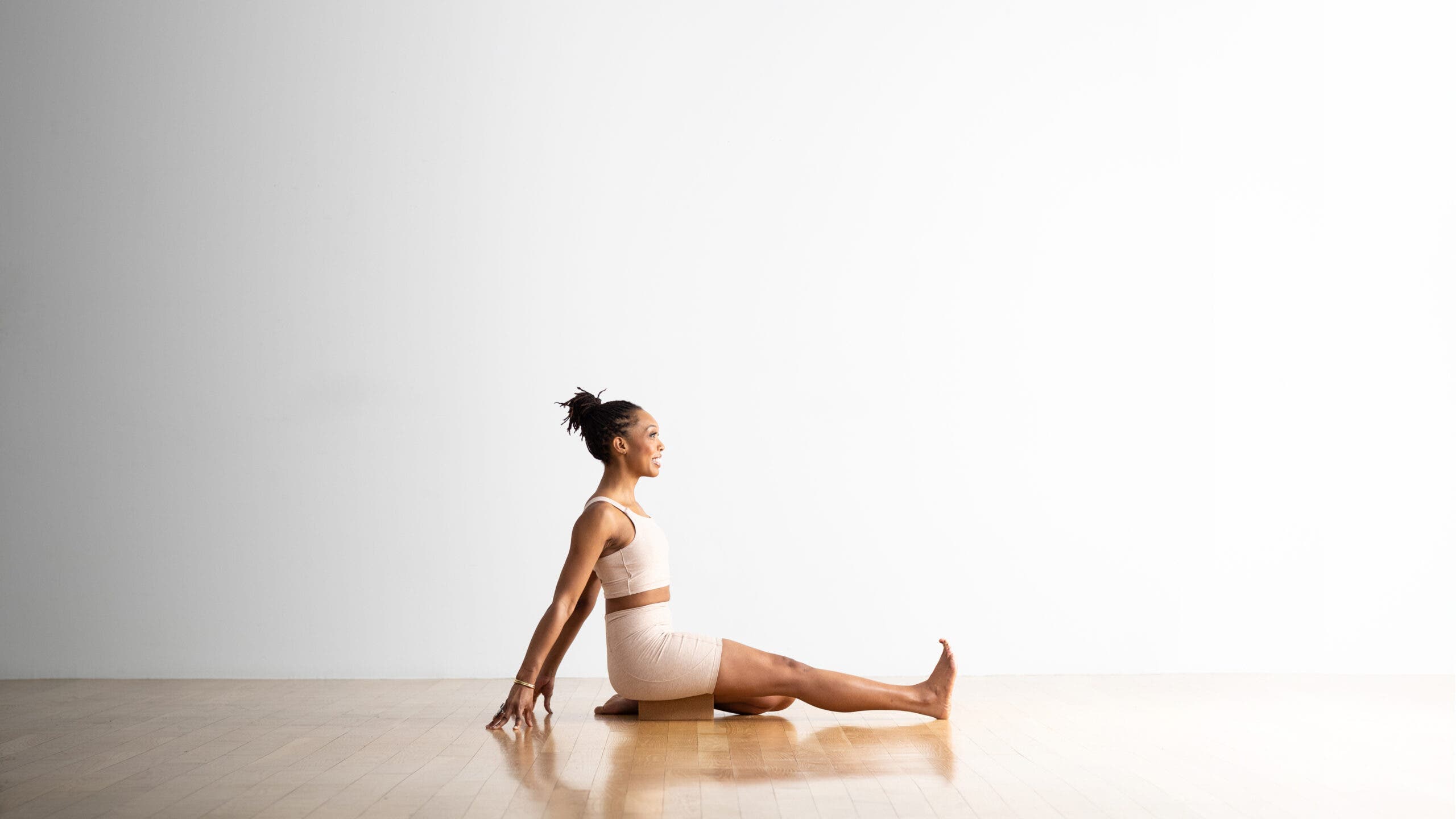Woman practices Supta Ardha Virasana (Half Reclining Hero Pose. She draw just her right leg back into Virasana, and the other leg is extended. She leans back and supports herself with her hands.. She is a Black woman with a ponytail, wearing off whit yoga shorts and a matching top. The floor is wood and the wall behind her is white.