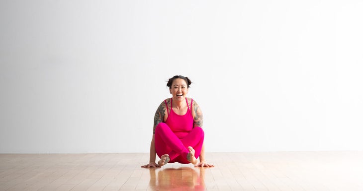 Soozie Kinstler practices Scale pose with legs crossed in Easy Seat. She is laughing, wearing bright magenta yoga tights and top.