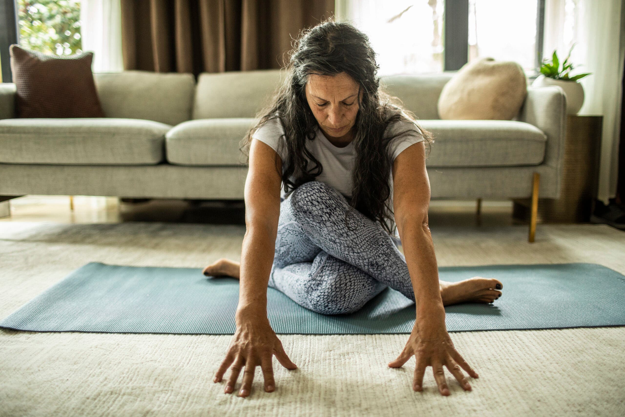 A woman with long salt-and-pepper hair leans forward and supports herself with extended arms. Her legs are crossed in Cow Face Pose. She is sitting on a blue yoga mat on off-white carpet. Behind her is a long oatmeal colored divan with brass legs. There's a brown pillow to match a brown curtain. You can see greenery in the sunny window behind her..