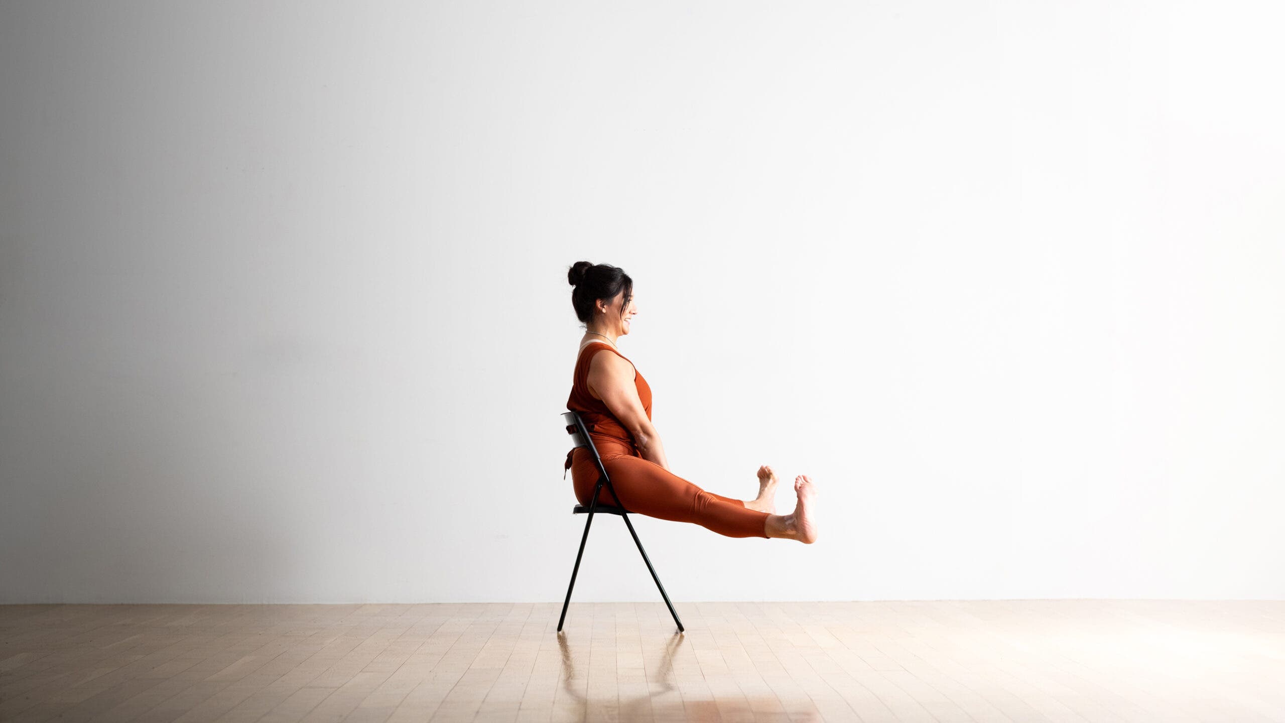 A yoga teacher practices Wide-Legged Seated Forward Bend sitting on a chair. She is wearing copper colored yoga tights and sleeveless shirt that ties in the back. The room has a wood floor and a white wall behind her.