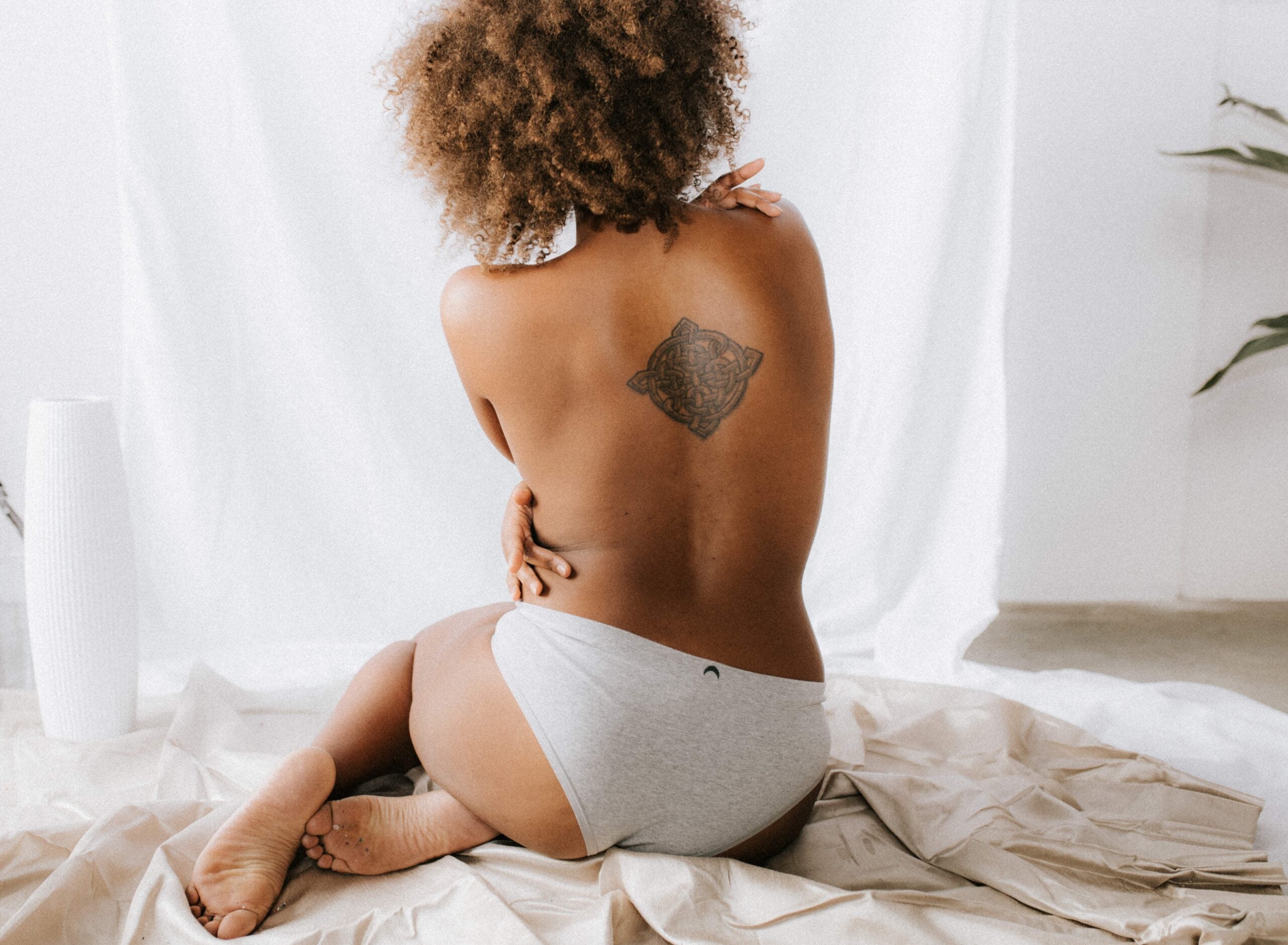 Black woman with curly hair sits on white sheets with her back exposed She has a tattoo between her shoulder blades