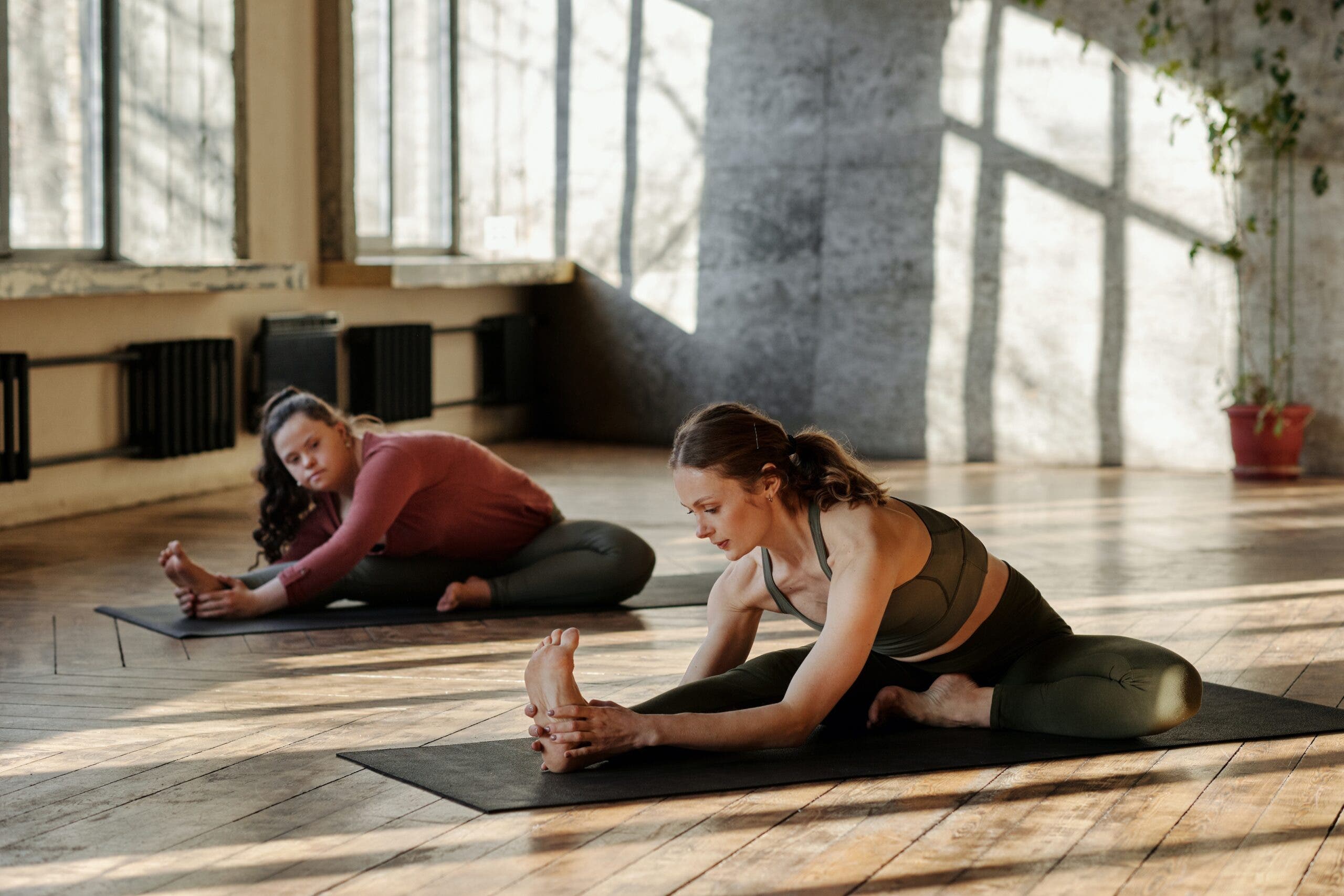 Two women practice Janu Sirsasana in a light filled industrial studio.