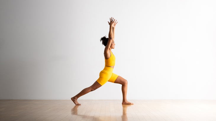 A brown-skinned woman wearing a bright yellow top and shorts, practices High Lunge with her arms extended up