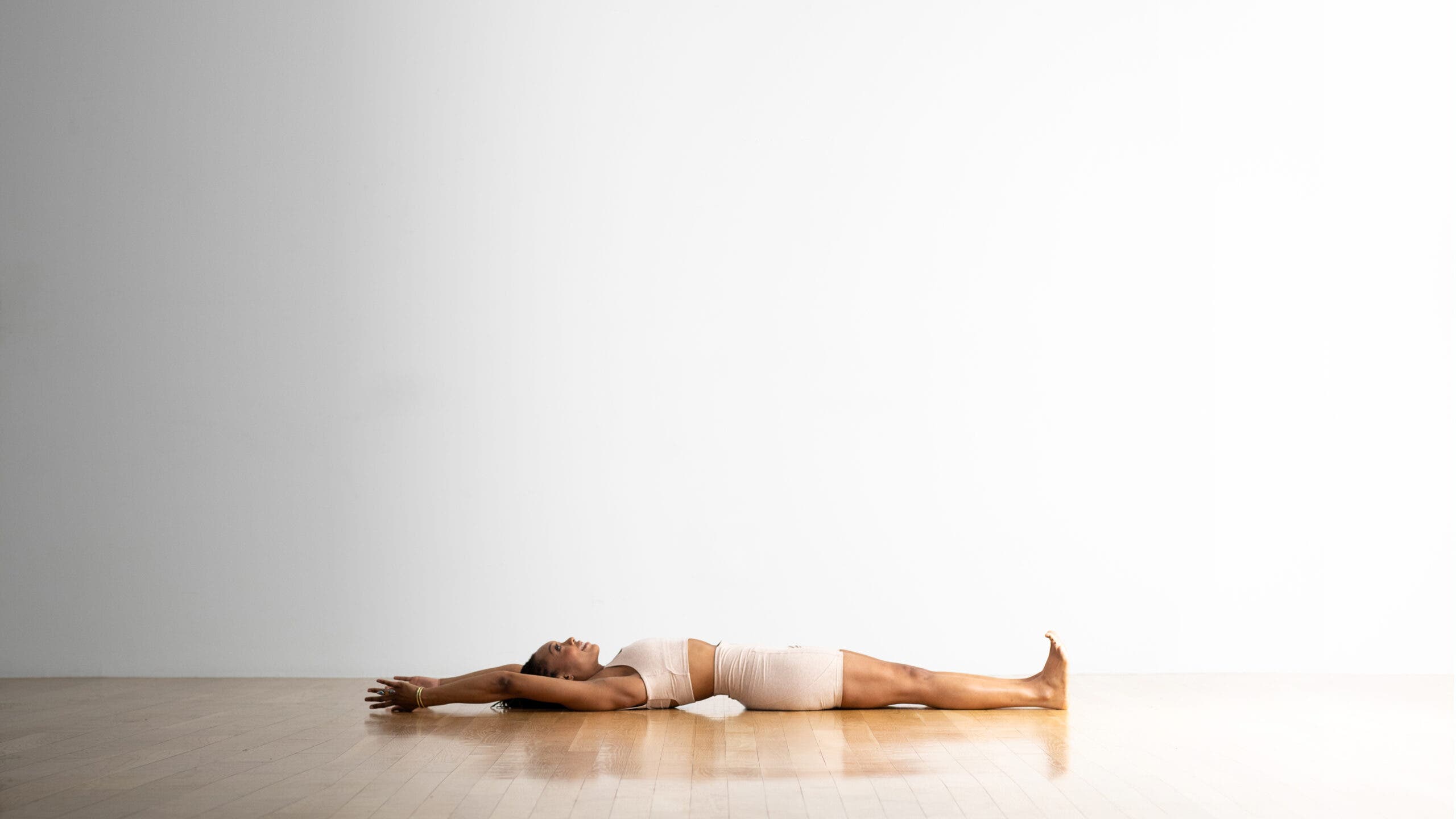 A woman practices Urdhva Hastasana lying on the floor. She is reclined on the floor with her legs extended and her arms reaching up over head.
