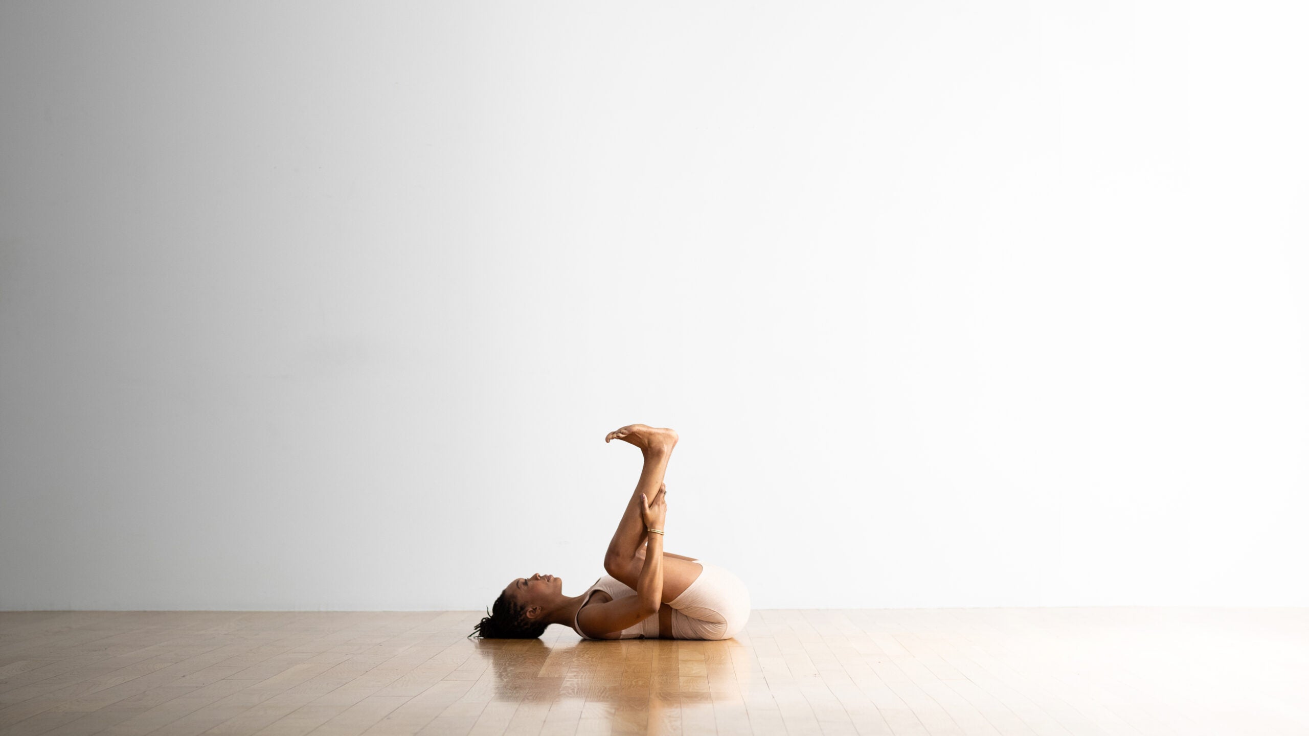 A yoga teacher practices a variation of Happy Baby Pose by putting her hands behind her calves instead of gripping the outside edge of her feet. She is a Black woman wearing light colored yoga shorts and a matching top. She is in a white room with a wood floor.