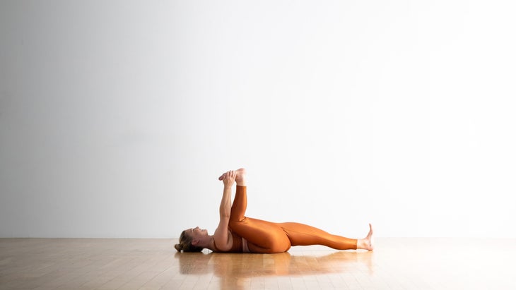 A woman wearing copper colored yoga tights and a matching shirt, practices a "half baby" pose--Happy Baby with one leg. She is in a white room with a wood floor.