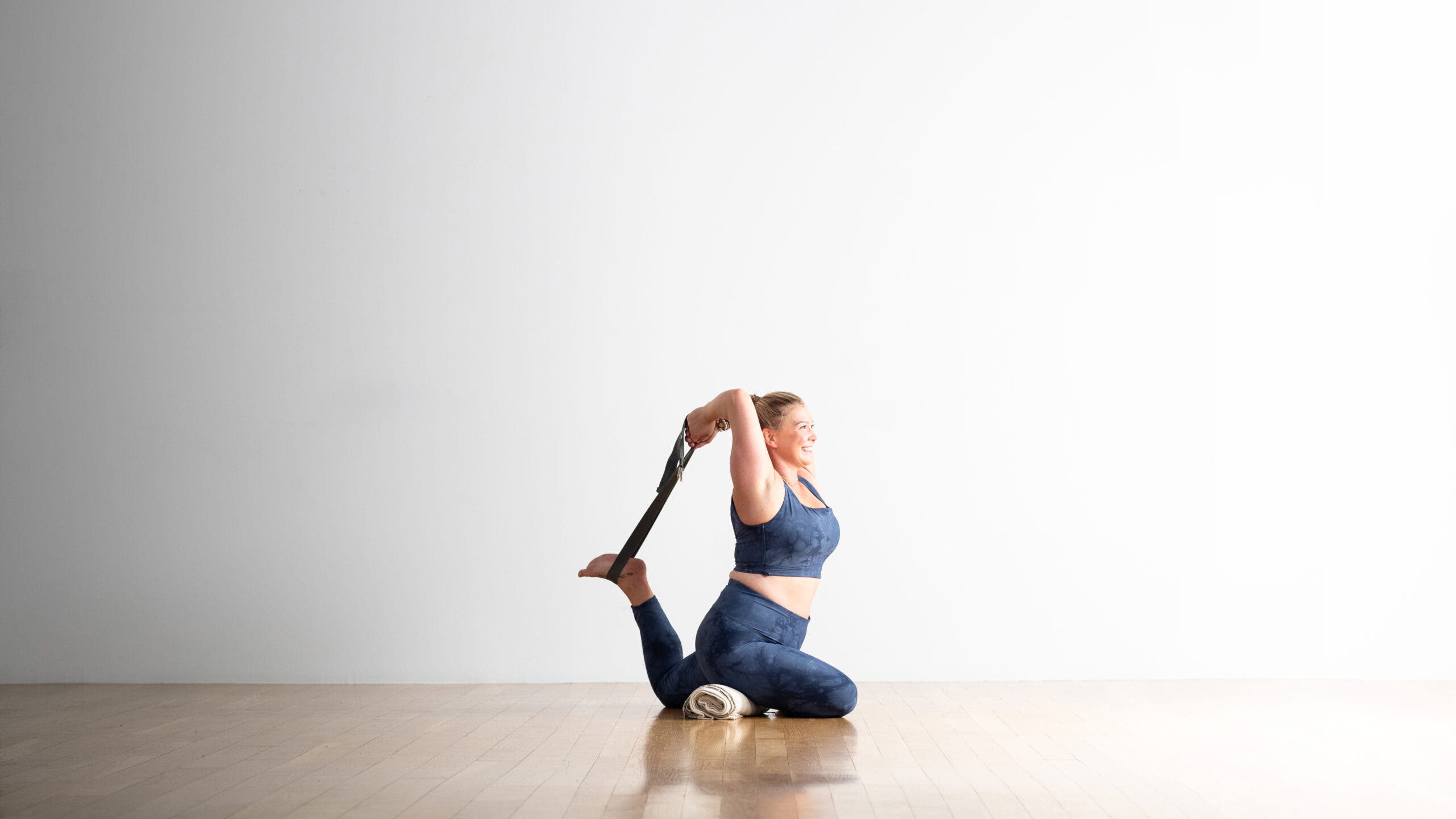 A woman practices One-Legged King Pigeon Pose with a strap around her back foot. She has a rolled blanket under her hip.
