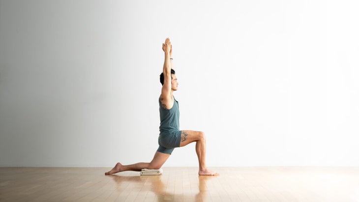 Man practices Low Lunge with his back knee on a blanket and his arms raised. He is wearing a gray-blue shorts and a sleeveless top. He has a tattoo on his shoulder and his thigh. The floor is wood and the wall behind him is white.