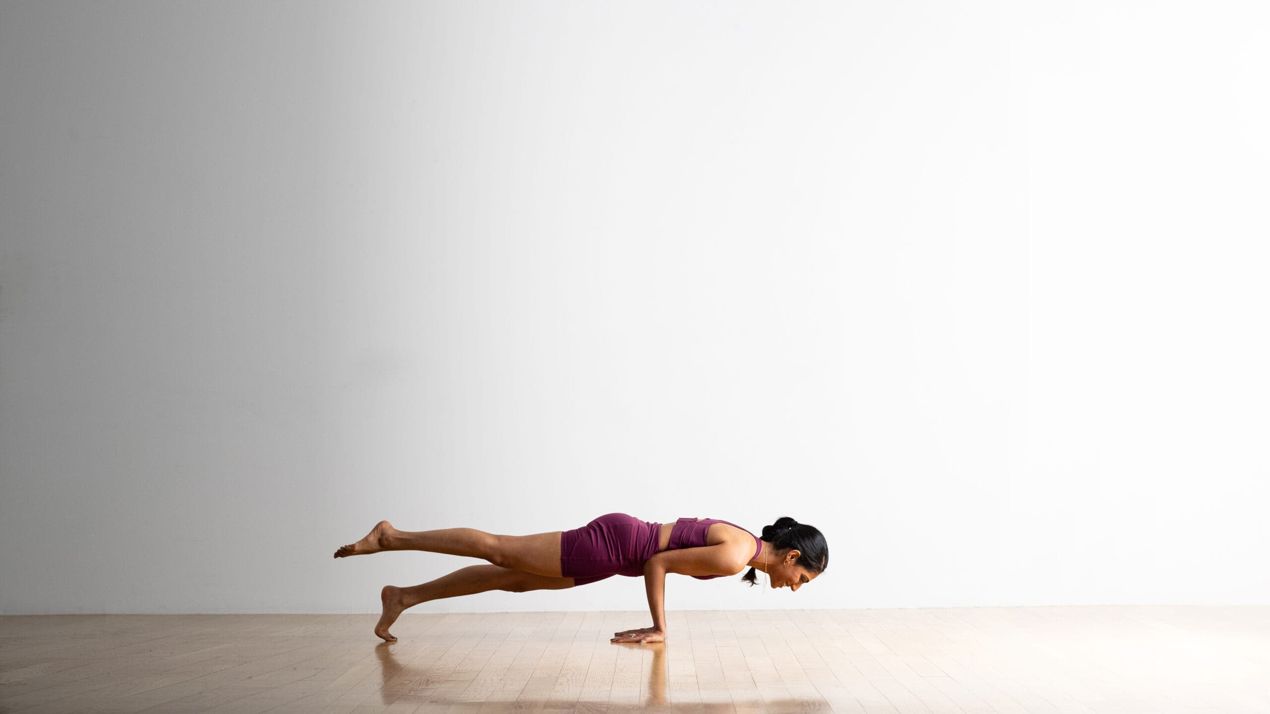A woman practices Peacock Pose with one foot lifted. She is a South Asian woman with dark ponytail. She is wearing purple shorts and a matching top. A white wall is in the background