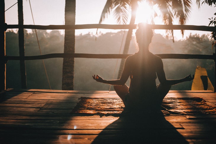 Slihouette of a person sitting on a balcony facing the sunset. She is in Lotus pose.