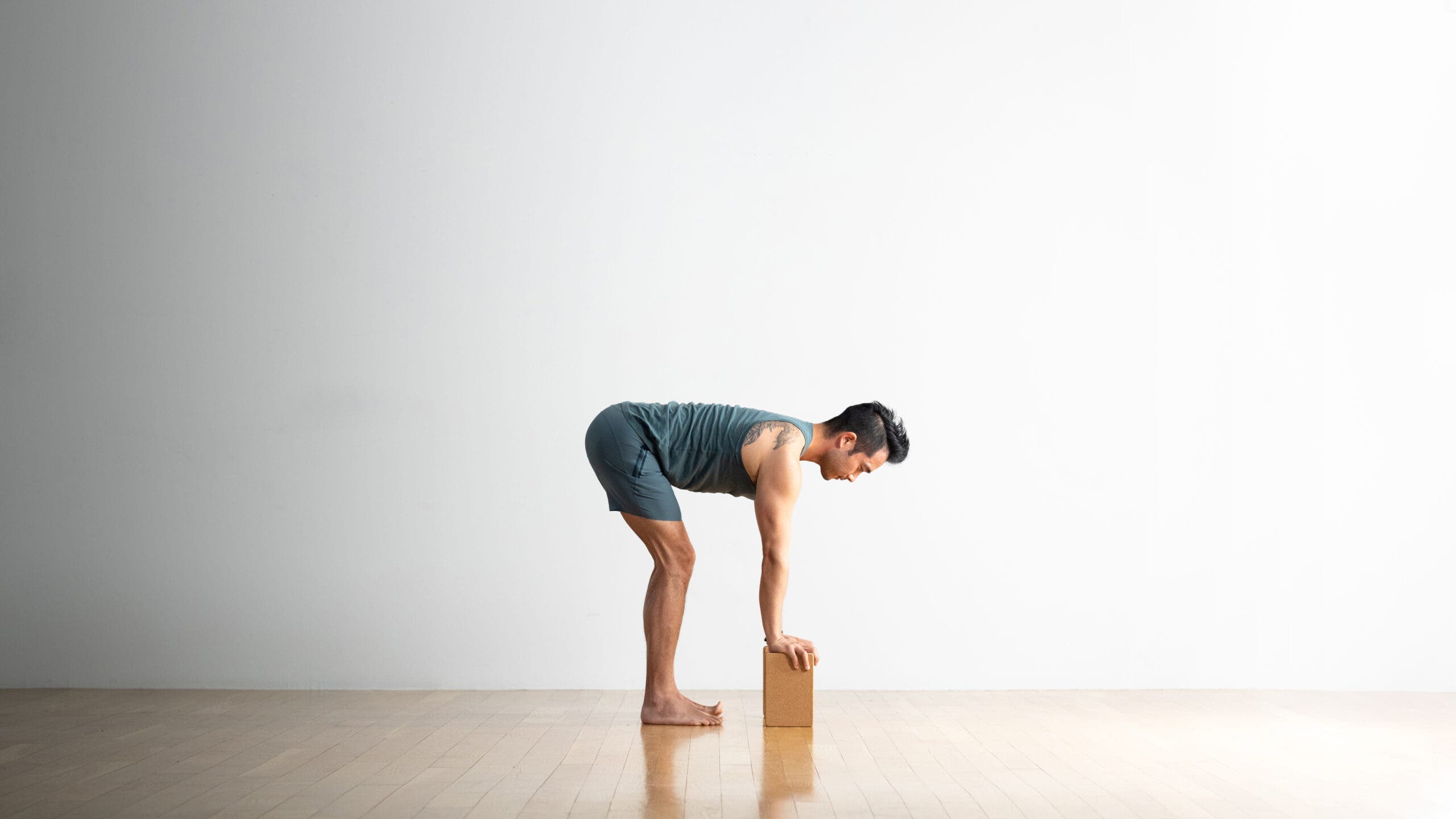 Man practicing Ardha Uttanasana (Standing Half Forward Bend) with cork blocks under his hands. He's wearing blue shorts and a sleeveless top. He has black hair and tattoos on his back and thigh.