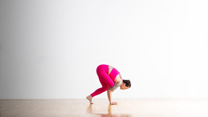 Soozie Kinstler practices a prep for Flying Pigeon Pose. From standing Figure Four pose she squats and puts her hands on the floor, then props her leg on her upper arm. She is wearing bright magenta yoga clothes.