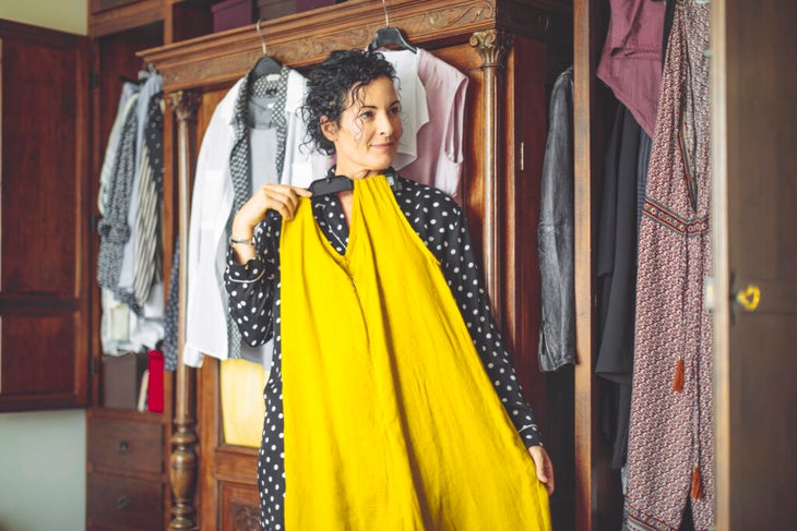 Woman holding up yellow dress to her body standing in front of closet.