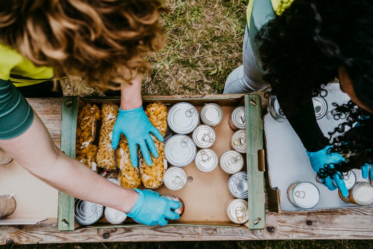 Two people reaching into box of canned food items.