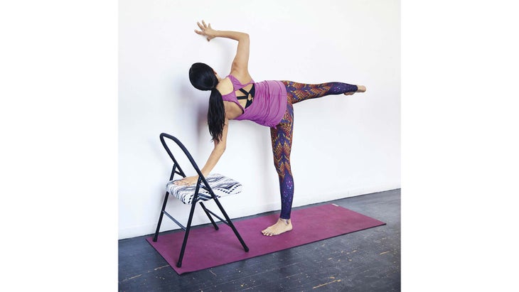 Person with long brown hair practices Half Moon pose. She is using a folding chair and a white wall as support.