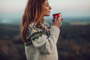 Woman with red hair in a sweater holding a mug of coffee and standing against a mountain background.