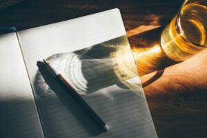 An open journal, a pen, and a yellow glass of water sitting on a wooden table in the sunlight.