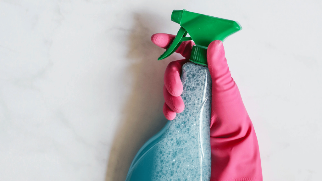 Hand wearing a pink disposable glove holding a spray bottle containing cleaning solution to kill dirt and bacteria