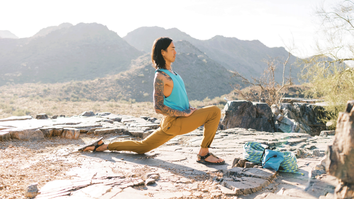 Man practicing yoga for climbers on a rocky ledge amid mountains by doing a low lunge with his back knee down and his hands on his front thigh