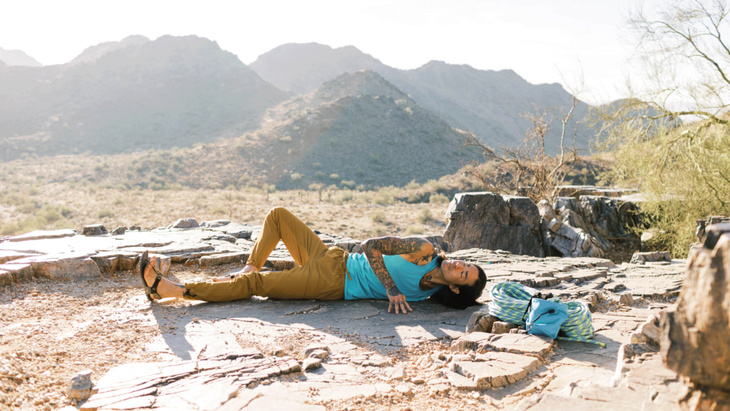 A rock climber lying on his side and stretching his shoulder and hip flexors on a rocky ledge in the desert with the morning sun backlighting him.