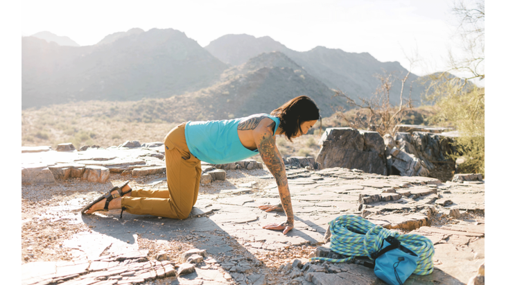 Rock climber on hands and knees practicing a stretch for the forearms on a rocky mesa among mountains. His climbing gear including rope and chalk bag is nearby.