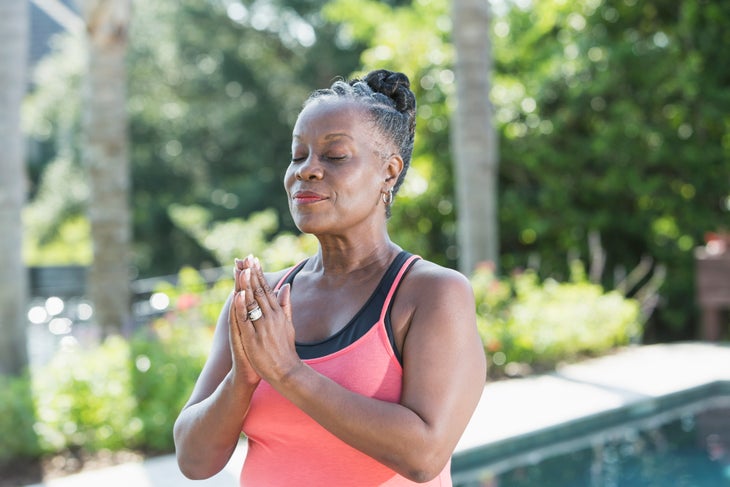 A mature African American woman standing outdoors with eyes closed, hands together. She is meditating, with peaceful, relaxed expression on her face