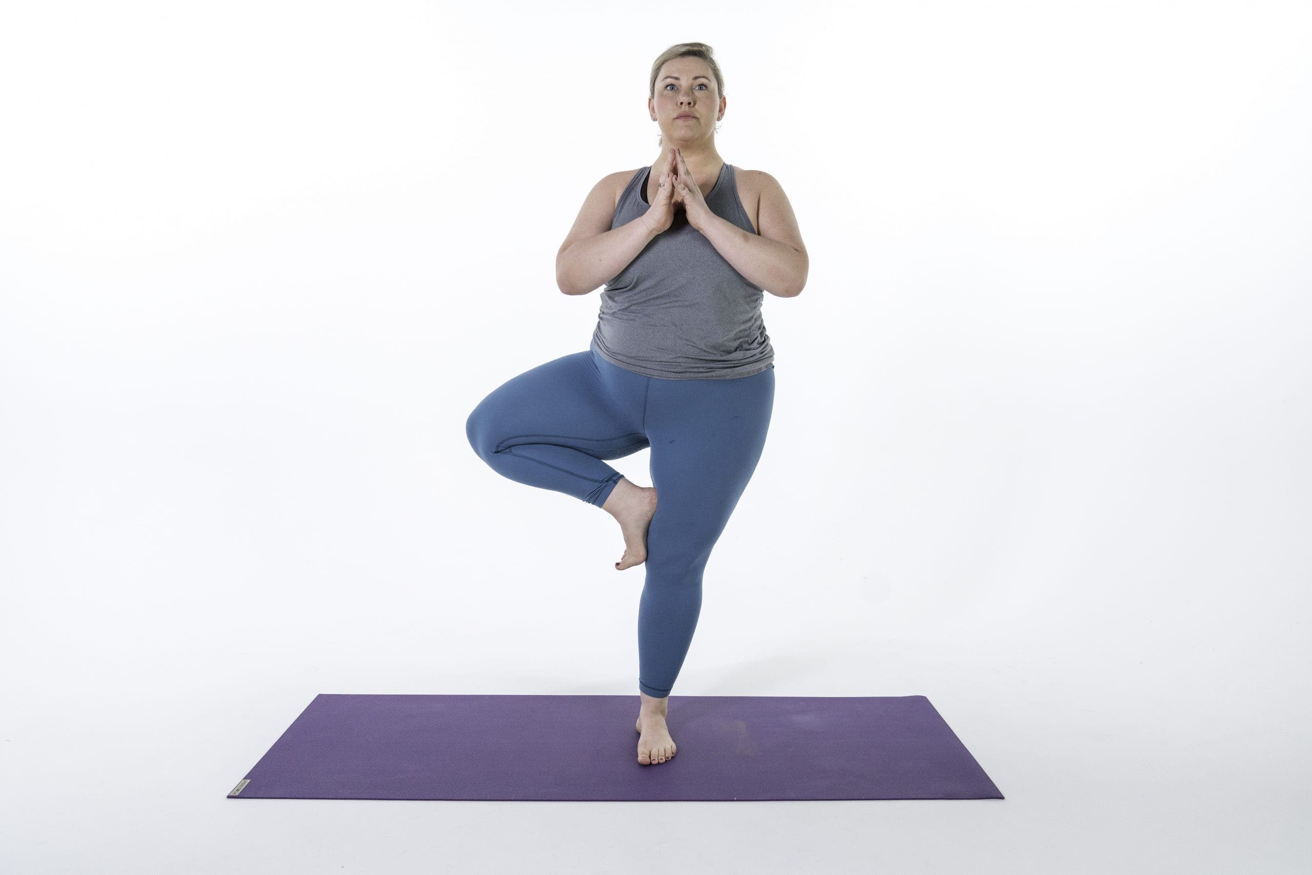 Curvy woman with blond hair, wears teal tights and a gray shirt while doing vrksasana_Tree Pose with hands in anjali mudra