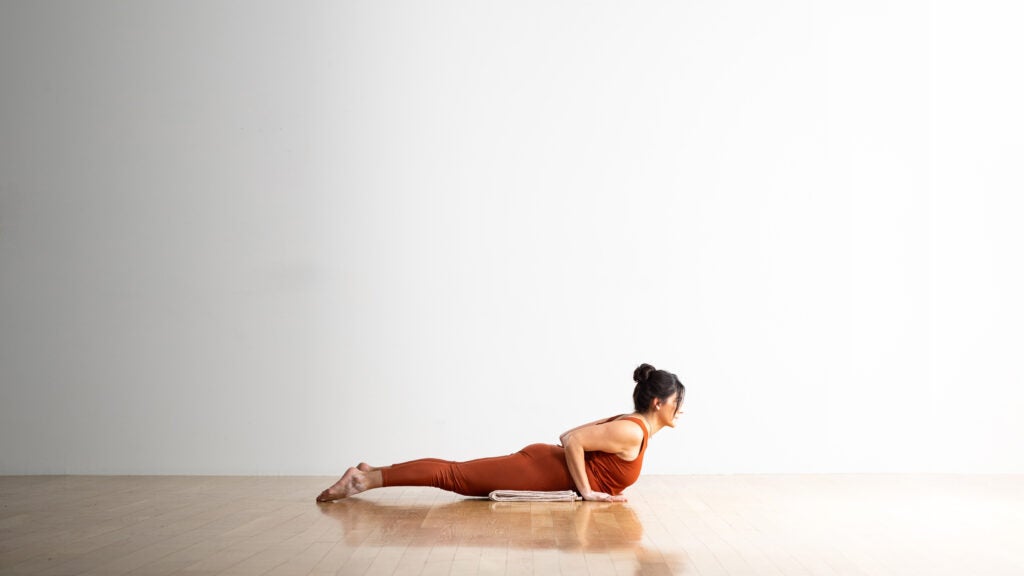 Woman with dark hair and copper-colored clothes practices Cobra pose. She is lying on a blanket placed on a wood floor. The wall behind her is white.