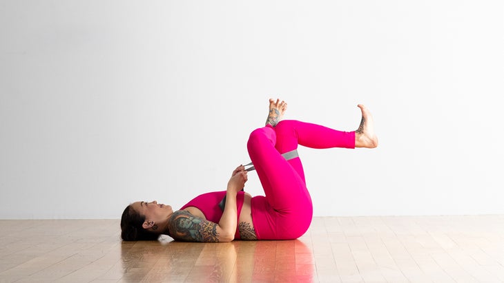 A woman practices a preparatory version of the pigeon pose.  She is lying on her back, both knees are raised to the body.  Her right ankle is crossed over her left knee, forming a quadrilateral.  She has a belt around her right hip and she holds it with both hands to pull it towards her.